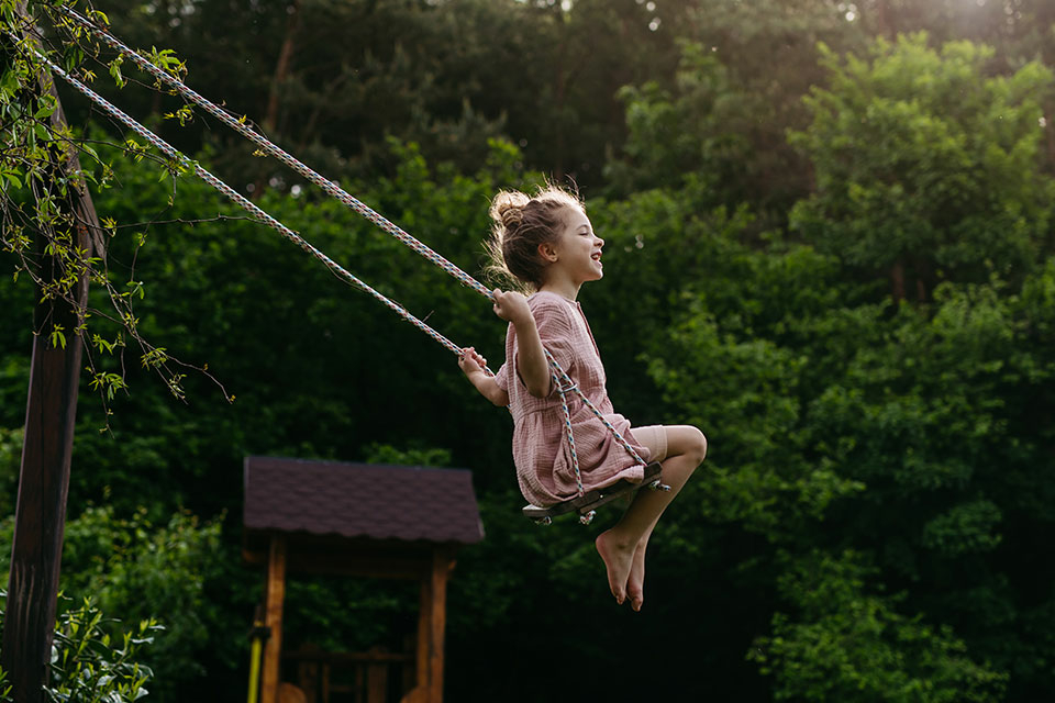 A young girl wearing a summer dress is swinging on a swing in a garden with trees in the background. 