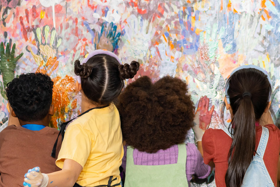 Six primary school-aged boys and girls from diverse backgrounds use their handprints to create a colorful abstract painting in a classroom. The children are different heights and are wearing colorful clothing. 